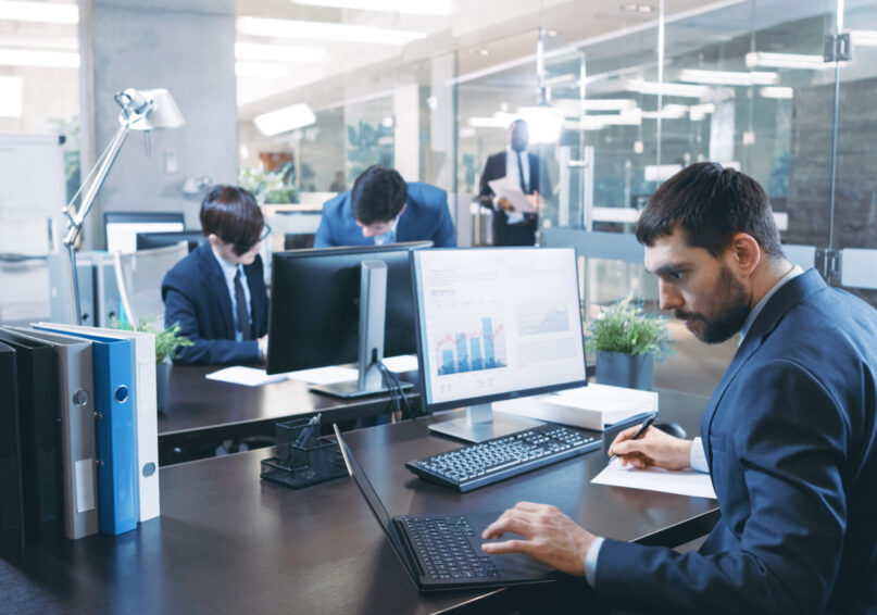 Professional Businessman Works on His Desktop Computer, Uses Laptop. In the Background Busy Office with Diverse Group of Business People.  Modern Glass and Marble Corporate Building.